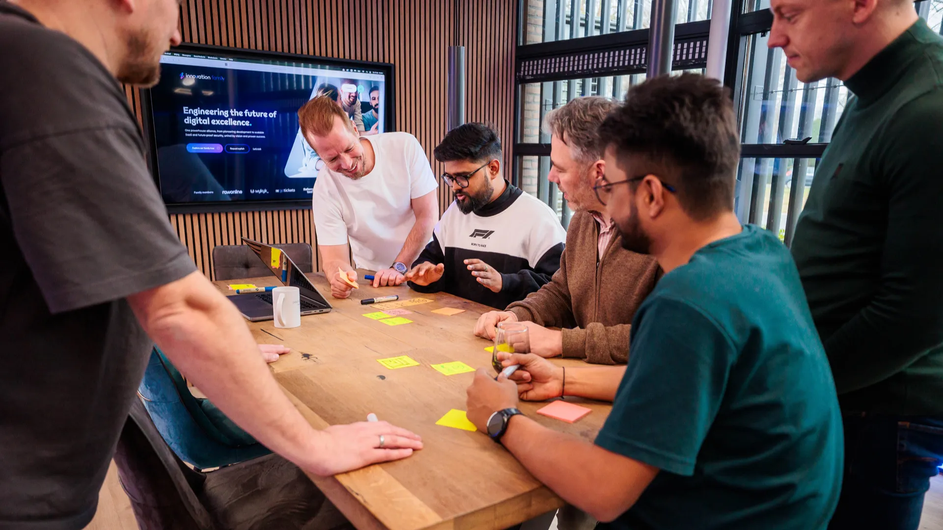 Zes teamleden staan rond een vergadertafel met post-its en markers tijdens een brainstormsessie, met een scherm met de Innovation Family website op de achtergrond.
